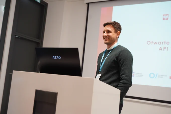 Na zdjęciu Maciej Wójtowicz stojący przy mównicy, na której leży laptop, wyświetlając prezentację na rzutniku, znajdującym się za jego plecami.
