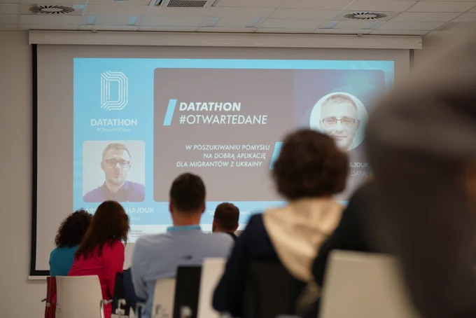 Na zdjęciu rzutnik z prezentacją online w sali z uczestnikami. Na rzutniku wyświetla się Arkadiusz Hajduk, tytuł prezentacji oraz logotyp Datathon #OtwarteDane