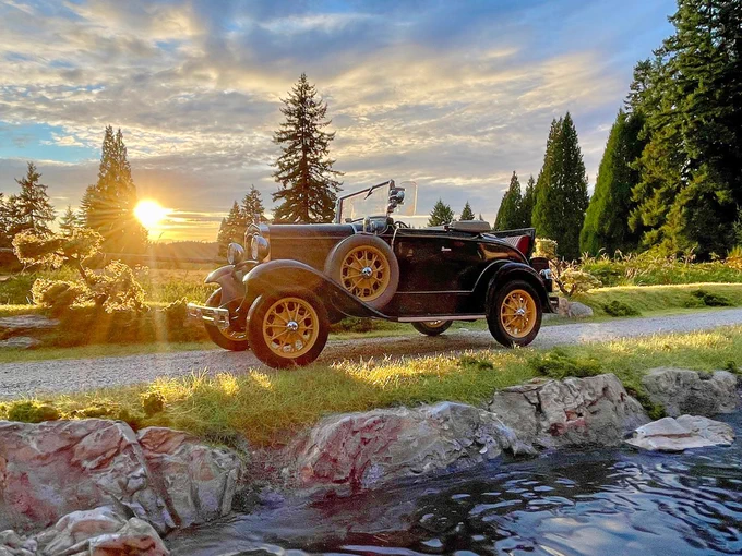Photo of a small, black, old-time model car parked on a miniature roadway situated behind a small flowing river. The car appears life-sized and the sunset is seen in the distance peeking over a field of green.