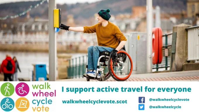 Man in wheelchair using pedestrian crossing with 'I support active travel for everyone' slogan