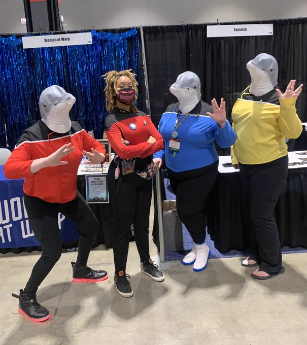 Aliza Pearl in Star Trek TNG uniform, posing with three cosplayers in Star Trek Lower Decks uniforms and dolphin heads, as Cetacean Ops. The 4 people stand in front of the Women at Warp and Fametek booths at Star Trek Mission Chicago.