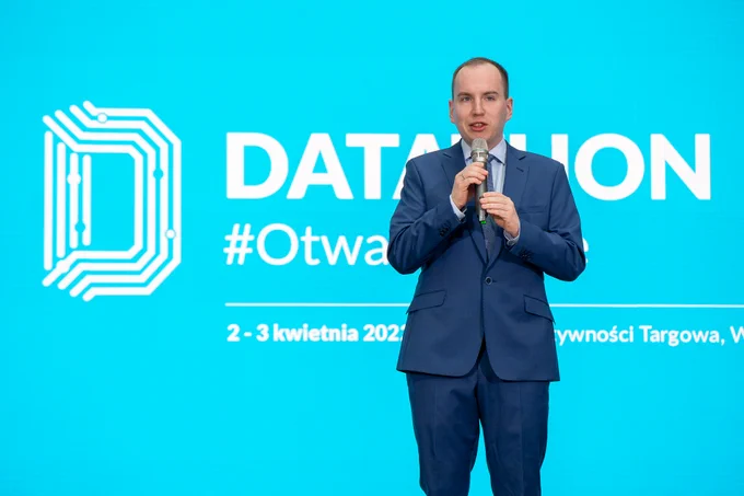 Na zdjęciu Adam Andruszkiewicz z mikrofonem na tle logotypu Datathon #OtwarteDane