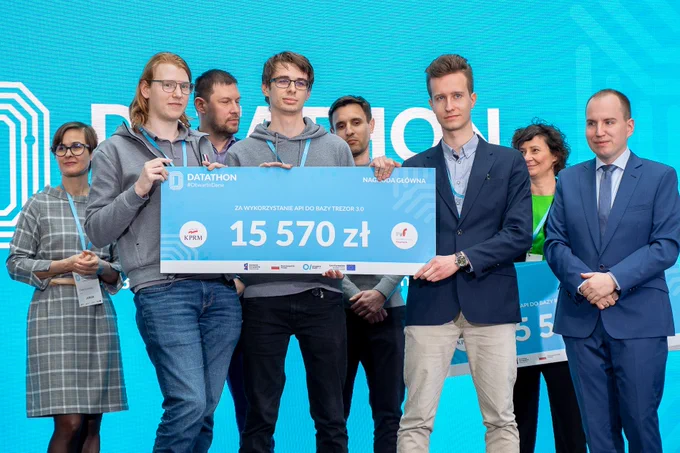 Na zdjęciu 8 osób, w tym zwycięski zespół Datathonu #OtwarteDane z czekiem w wysokości 15 570 PLN wraz z Ministrem Adamem Andruszkiewiczem.