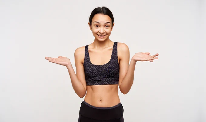 young woman similing and shurgging her shoulder whilst wearing a black sports bra standing in front of a pale grey background