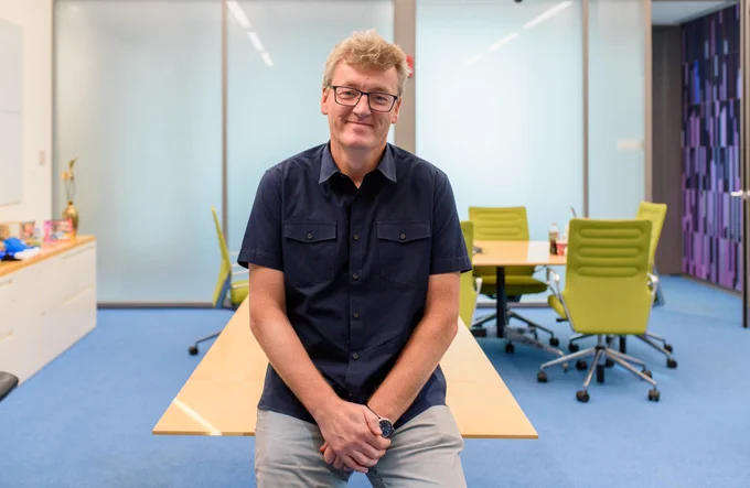 Dr. David MacMillan, wearing a blue button up shirt and grey pants, sits on a desk.