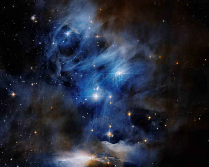 Composite image of a nebula, with wispy blue clouds of dust and gas. Interspersed are stars, some brighter than others. Toward the center of the image are two seemingly large, bright stars. Pockets of void are in the image, showing the blackness of space. 