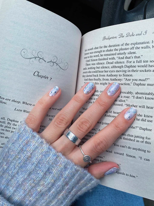 Hand painted pastel marbled pink, white, baby blue and lavender nail design with three gemstones on each nail on a left hand place on a page of the book “The Duke and I” by Julia Quinn book one of the Bridgerton series.