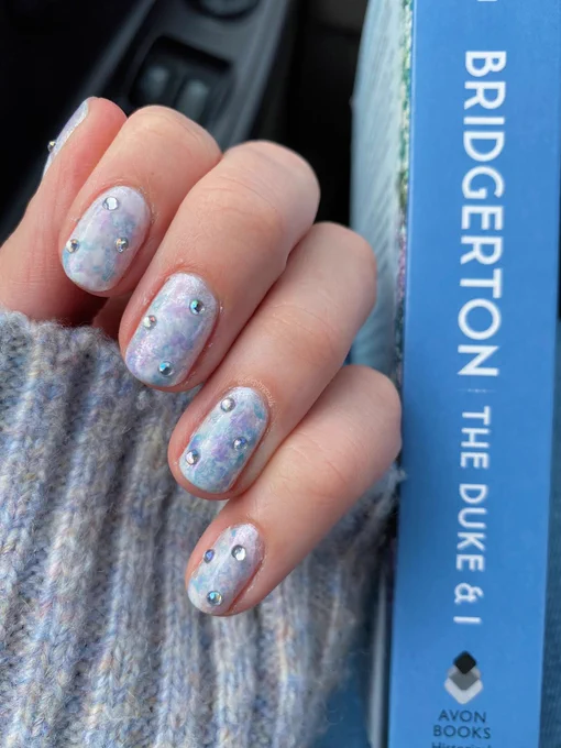 Hand painted pastel marbled pink, white, baby blue and lavender nail design with three gemstones on each nail on a left hand in a fist position next to the spine of the book “The Duke and I” by Julia Quinn book one of the Bridgerton series.
