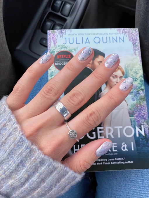 Hand painted pastel marbled pink, white, baby blue and lavender nail design with three gemstones on each nail on a left hand in front of the book “The Duke and I” by Julia Quinn book one of the Bridgerton series.
