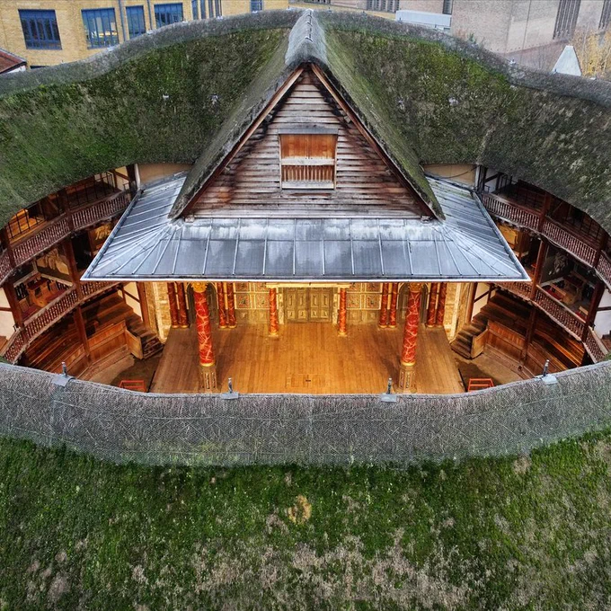 A aerial shot looking into the Globe Theatre from above, the thatched roof is littered with green moss and the wooden thrust stage is cast in a golden warm glow.