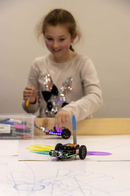 Your girl excited to watch as her robot drives across paper sheets on a robotics table. The robot has a pen attached and is drawing.