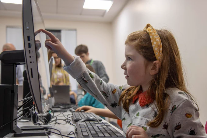 Young girl using a touch screen computer to create a program. Other children coding in the background.
