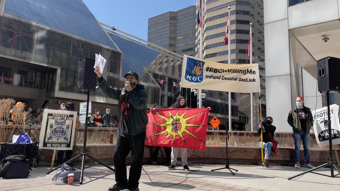 An Indigenous land defender is pictured speaking at the Toronto RBC action in front of banners that say “#RBCIsKillingMe” Defund Coastal GasLink” and “Protect Wet’suwet’en Land. No Pipeline.”