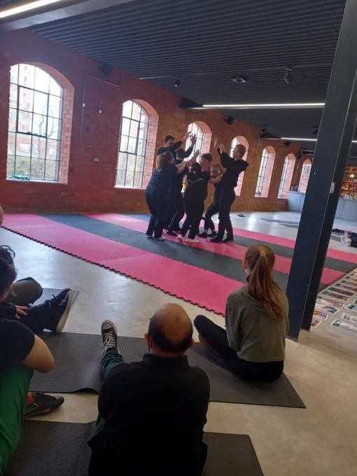 Hubbub Actors and facilitators making the shape of a tree on black and red mats in The Museum of Making. We can see the audience watching them as they are sat on the floor. 