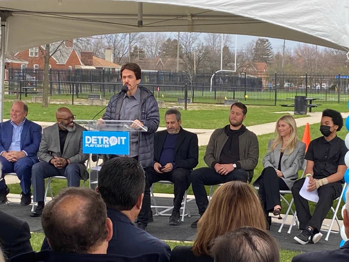 Mitch Albom speaks at a podium while seated behind him are Mayor Mike Duggan, SAY Play Executive Director Eric Reed, SAY Play COO Marc Rosenthal, Matthew and Kelly Stafford, and a student from SAY Play. 