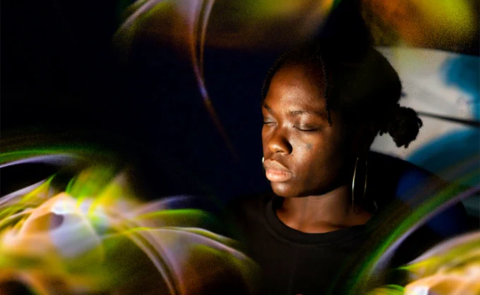 A woman sits in a dark room, a beam of light illuminates her face as swirls of light circle her.