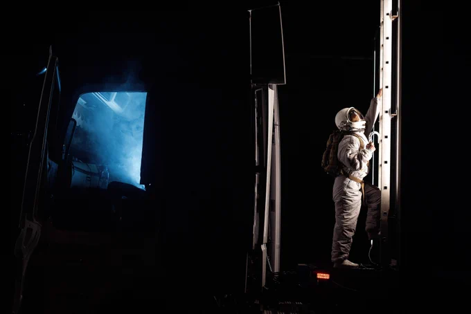 An astronaut reaches up a ladder as it climbs up the side of a container on the back of a lorry. The cab of the lorry is open and blue smoke billows out. It is night-time, the only light a blue beam coming out of the cab and a bright light illuminating the astronaut.