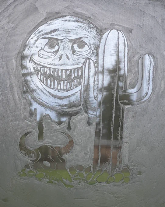 a silk screen is held against a window. the background is a few blurry trees with grey/blue skies peaking through. on the silkscreen is glue in the shape of a cactus, with some rocks at the bottom and a cow skull next to it. along with the image of a really creepy looking sun. it has a face that is smiling but looks sinister in the eyes. it has baggy eyes and crows feet and the teeth are thin and decaying. it's nose is snake-like and it is also dripping something I didn't honestly know what but it felt like it needed drips. also the sky in the background is shining through the sun and mostly just the sun. trees fill out the rest