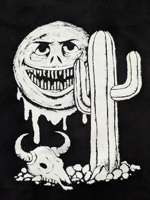 a patch freshly printed on black fabric. it is printed with white ink and shows a three pointed cactus and a cow skull surrounded by rocks in the foreground. looming behind it is a very creepy looking sun with a graveyard of uneven, thin teeth. it appears to be smiling, but it's brows are also furrowed and pointy, it has a nose like a snake and thin lips stretching out on both sides. it is also oozing or sweating in large drops. there's tons of texture of the face of the sun in a big swirl pattern. there's little bits of textures on the cactus and cow skull to add shadow as well