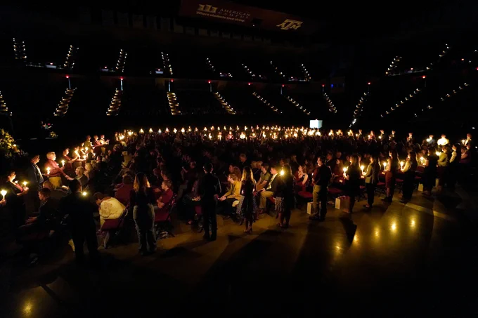 Reed Arena in darkness as people are seated in the stands and in chairs on the floor as hundreds of Aggies stand with candles surrounding the Aggies on the floor of the arena