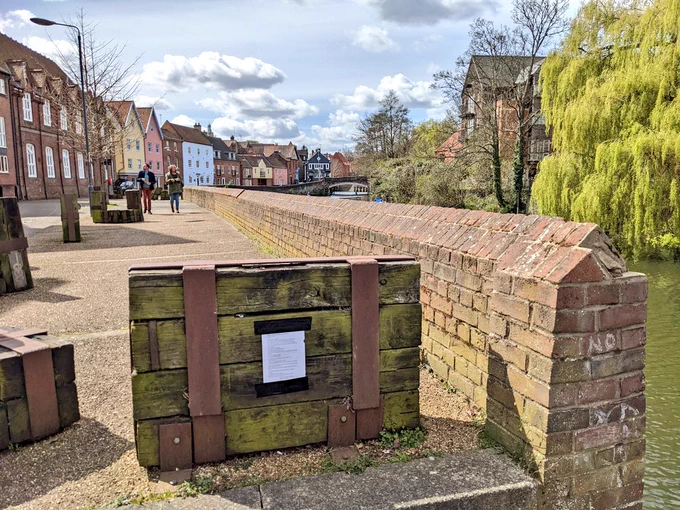 A photo of a riverside walk, featuring wooden art pieces reminiscent of ship cargo. Taped to one of these is a page of poetry.
