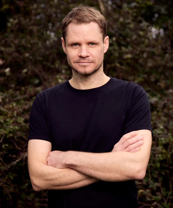 White person with light brown hair and a tiny bit of facial hair.

Wearing a black t-shirt. Muscly arms. 