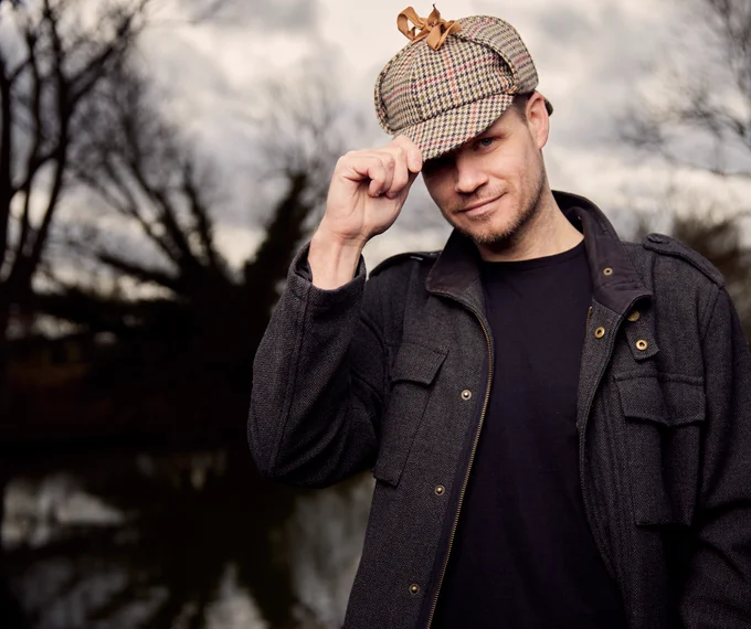 a white person with stubble, wearing a black jacket and a cap. The background is moody. Trees and twilight.