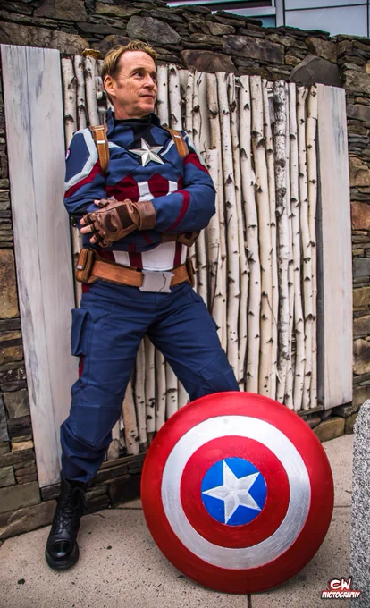 A man cosplaying as Captain America with arms crossed, leaning against a wall