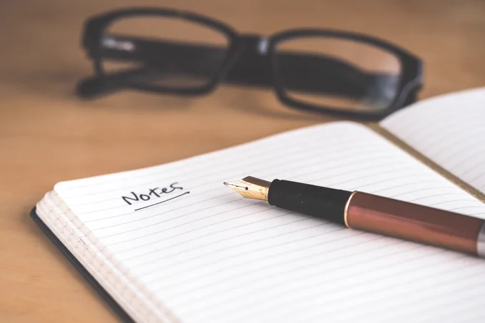 Open notebook with a fountain pen laying on top. "Notes" is written on the top of the page and underlined. A pair of black glasses sits in the background. Photo by David Travis on Unsplash