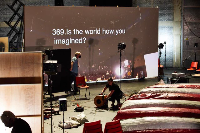 A big screen on a stage reads "369. Is this the world how you imagined?" In front three people are working on the stage. One has headphones on, another is working with cables.