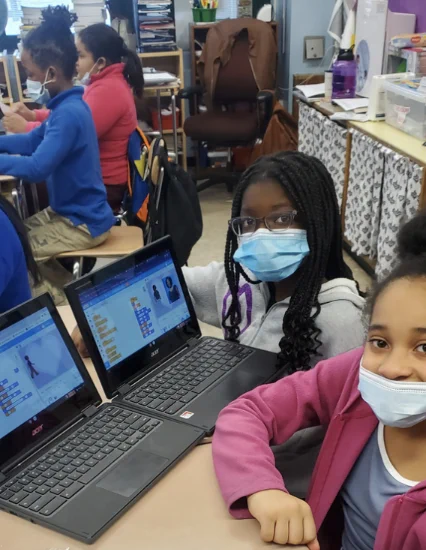 two girls wearing a mask showing projects on their laptops