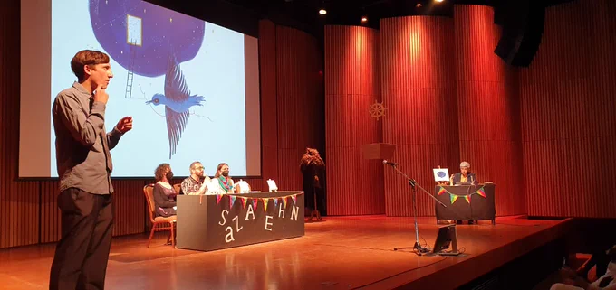Sobre escenario de un auditorio, En el centro hay un mesón con tres personas sentadas detrás de él. A la izquierda se aprecia a un intérprete de lengua de señas y en el fondo, a la derecha, se ve otra mesa con una persona detrás de ella. 