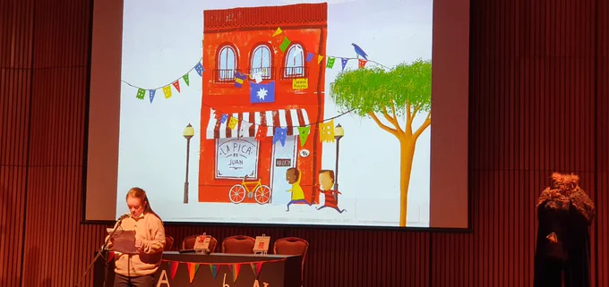Sobre escenario de un auditorio hay una joven con discapacidad intelectual leyendo frente a un micrófono. En el fondo se proyecta una ilustración de un negocio con dos niños corriendo en una calle. 