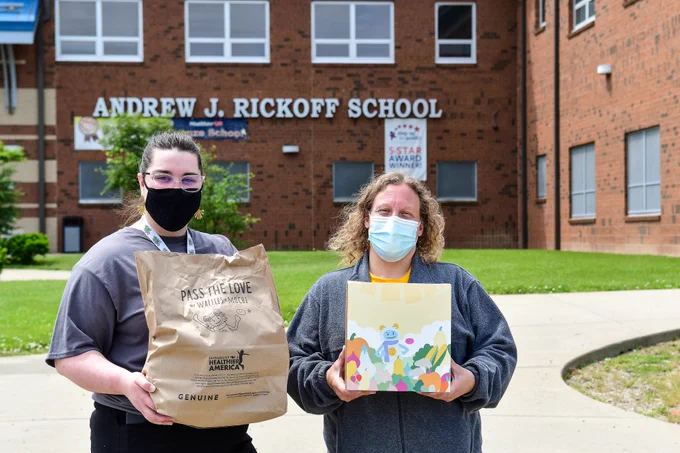 Photo of two volunteers holding a bag with Pass the Love meal kits in front of a school distribution site. 
