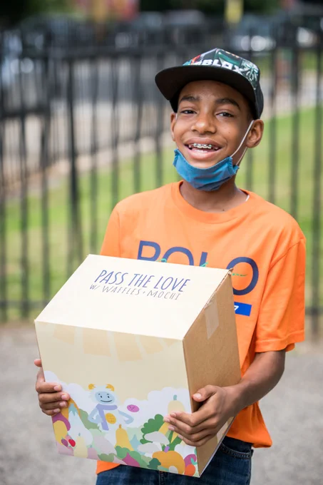 A young boy holds one of the Pass the Love meal kits featuring healthy food items. The box is decorated with images of the characters from the Netflix show, Waffles   Mochi. 