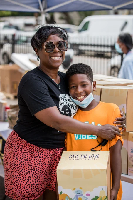 A mother and son hug while standing at a Pass the Love distribution site and holding a healthy meal kit. 