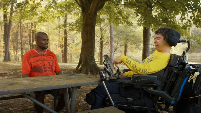 Andrew and Samuel sitting across from each other at a wood table. Andrew is wearing a red t-shirt that says "November Project" and Samuel is in his wheelchair, wearing a yellow long sleeve shirt that says "Go Blue" on the sleeve. There are many tall trees behind them with green leaves.