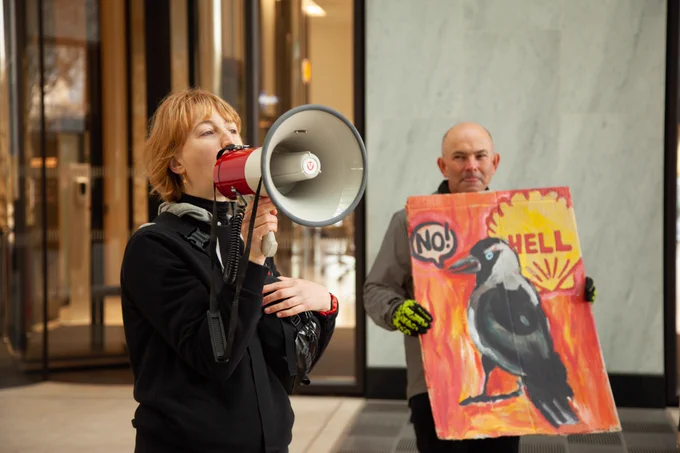A woman with fringe shouts through a red megaphone, a red Hell Shell placard with Jackdaw on it is held by a man in the background