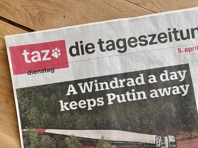 Foto der Titelseite der taz vom 5. April 2022. Die Zeitung liegt auf einem Holztisch. Auf der Titelseite ist ein Bild eines LKW zu sehen, der ein Rotorblatt eines Windrades transportiert. Er fährt durch einen Wald. Die Schlagzeile zu dem Bild lautet: A Windrad a day keeps Putin away.
