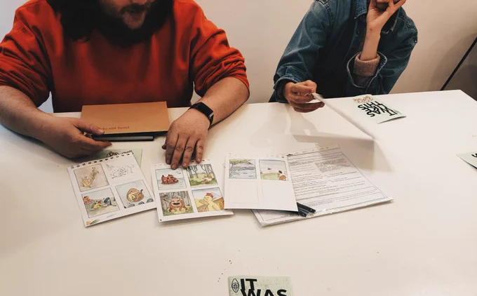 Two people sitting at a table looking through art works in progress 