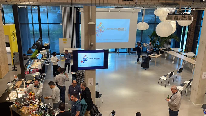 A group of people standing in front a buffet. Some more people distributed among a large space in groups, chatting. Two screens displaying the ServerlessDays Amsterdam logo