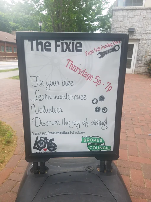 A rectangular sign on a sidewalk titled in bold font "The Fixie." In the top corner in diagonal red font reads "Eagle Hall parking garage" over a picture of a bolt wrench, followed by the text "Thursdays 5p - 7pm." Cursive text reads down the center "Fix your bike, Learn maintenance, Volunteer, Discover the joys of biking!" In smaller text at the bottom reads "Student run. Donations optional but welcome." The logos for the Emory Bike Social and Emory Spoke Council are in the bottom corners.  