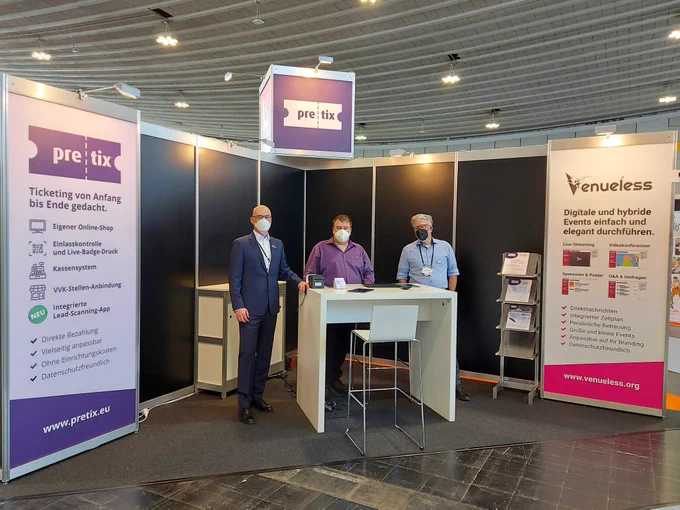 Unser Stand auf der BOE connect live. Links ist eine Wand mit Infos zu pretix bedruckt, rechts eine mit Infos zu Venueless. In der Mitte ist oben ein Kasten mit dem pretix Logo. Unten steht ein Tisch mit Infomaterial. Dabei stehen unsere Mitarbeiter Jochen Siebert, Martin Gross und Florian Bokor.