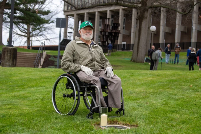 Dean Kahler, one of the nine students wounded on May 4, 1970, sits in a wheelchair in front of the plaque marking the location where he was wounded.