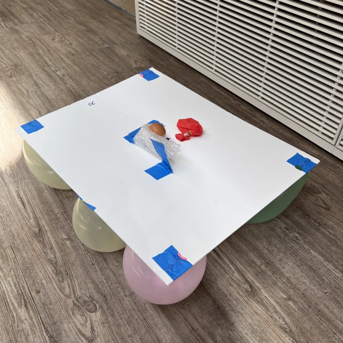 quatre ballons sous un carton assez grand et là, aux coins, un œuf dans un gobelet enveloppé de papier bulle, une anche fabriquée maison sur un ballon (ici vide)
