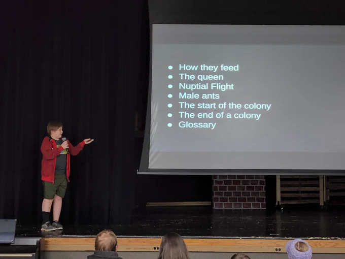 A student on stage, pointing to a slideshow screen, featuring bulletted text about ants.