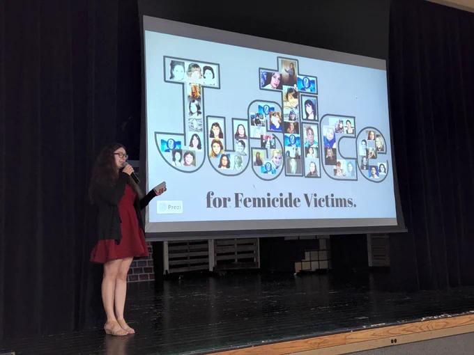 A student on stage, pointing to a slideshow screen with the word "Justice", filled in with photos of female murder victims.