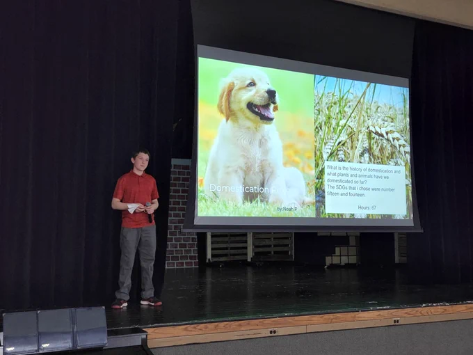 A student on stage, next to a slideshow screen featuring a golden retriever puppy, stalks of wheat, and some text.