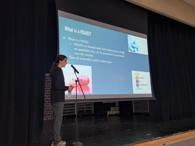 A student on stage, with to a slideshow screen featuring text and photos about painkillers.