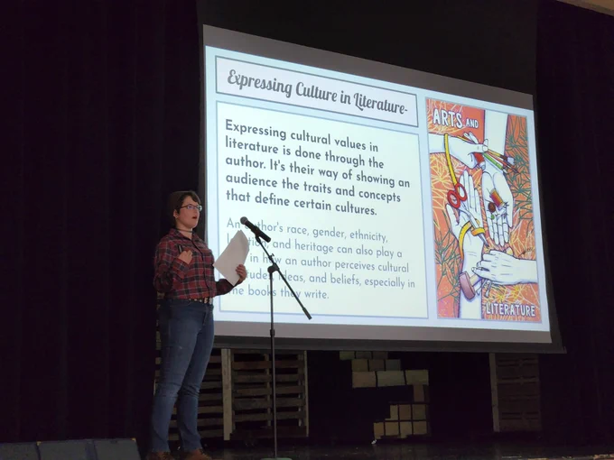 A student on stage, next to a slideshow screen with text and graphics about literature and culture.